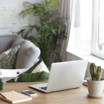 Stylish home office with laptop, cactus, smartphone, and green smoothie on wooden desk near sofa and large window