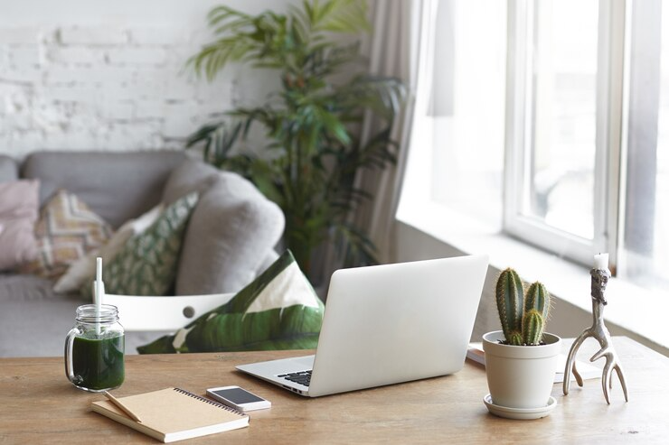 Stylish home office with laptop, cactus, smartphone, and green smoothie on wooden desk near sofa and large window