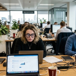 Office workspace with diverse employees focused on laptops, modern open-plan design, plants and city view in background